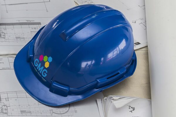 A blue hardhat with the GMG logo sits on a table on top of some blueprints